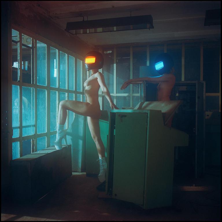 Two nude women wearing helmets with illuminated visors pose in a room with large windows and industrial equipment.