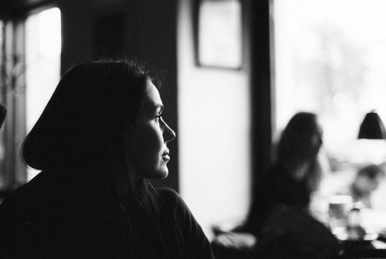 A woman is seen in profile, looking out a window, with another person out of focus in the background.