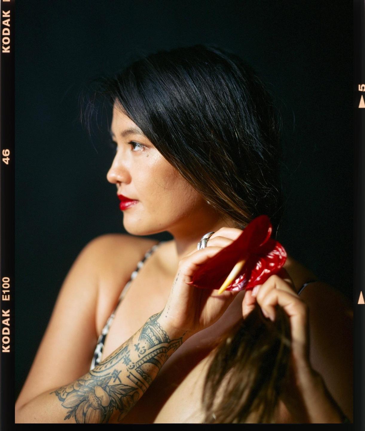 A woman with long dark hair and red lipstick holds a red anthurium flower.