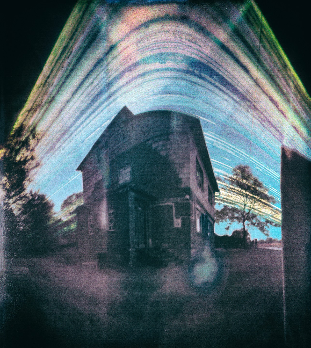 An abandoned house stands under a sky with light trails.