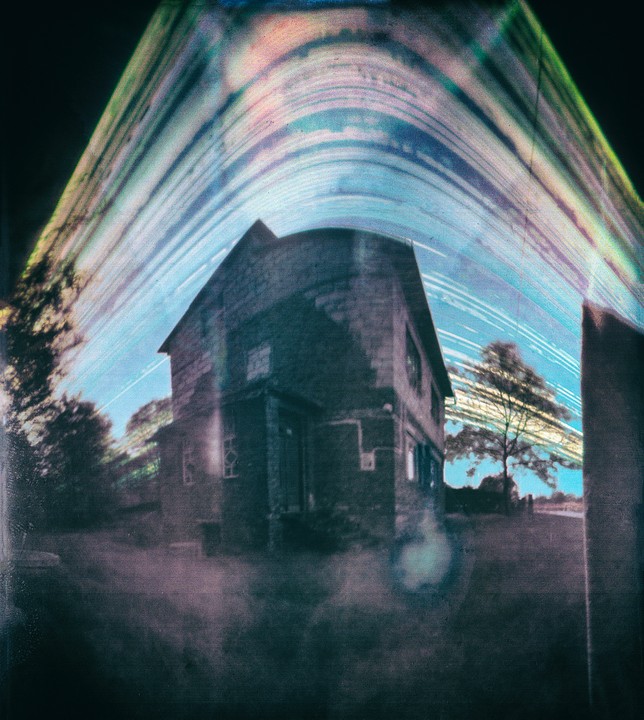An abandoned house stands under a sky with light trails.
