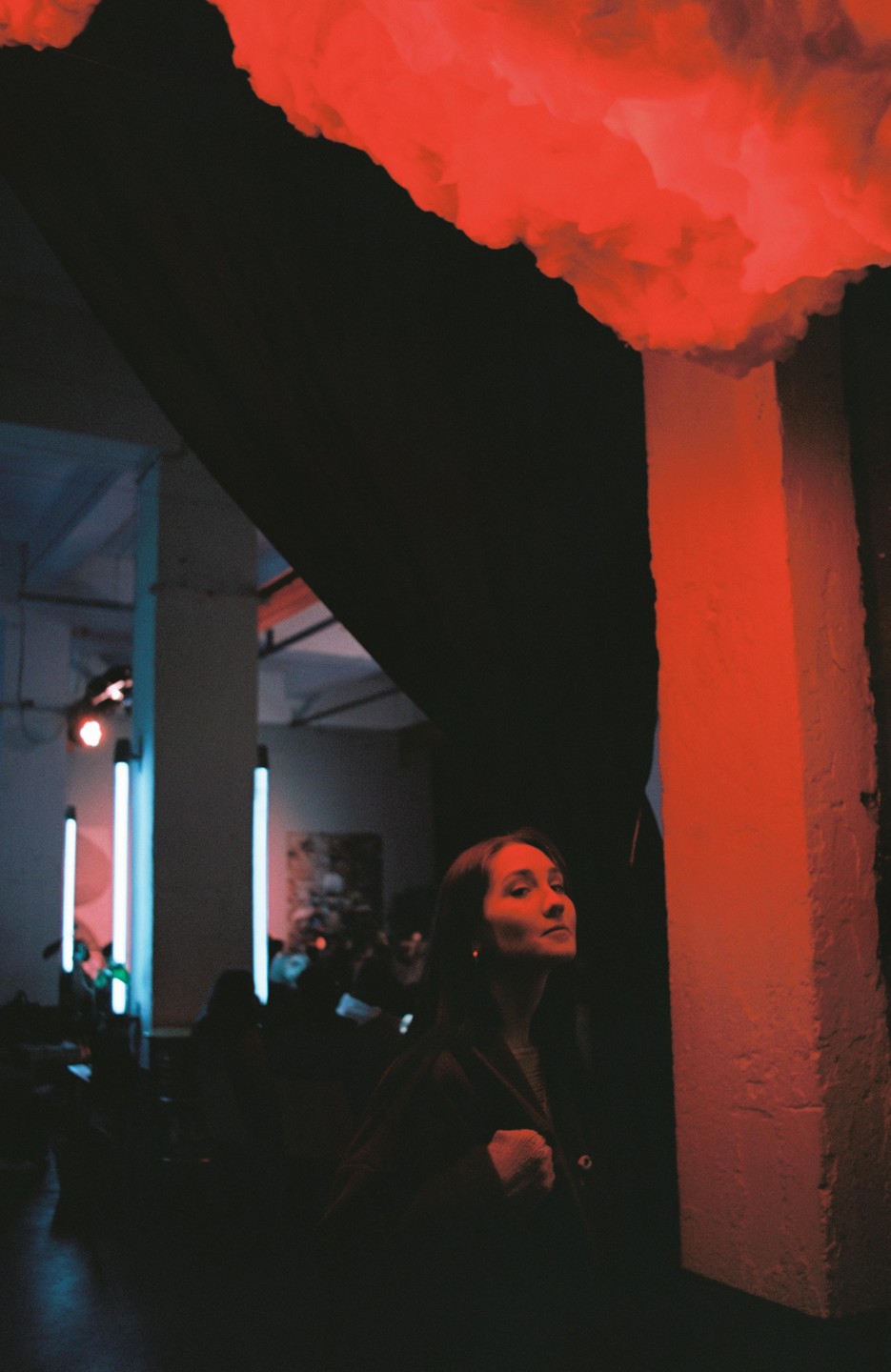 A woman looks up at a red cloud installation in a dark room with blue neon lights in the background.