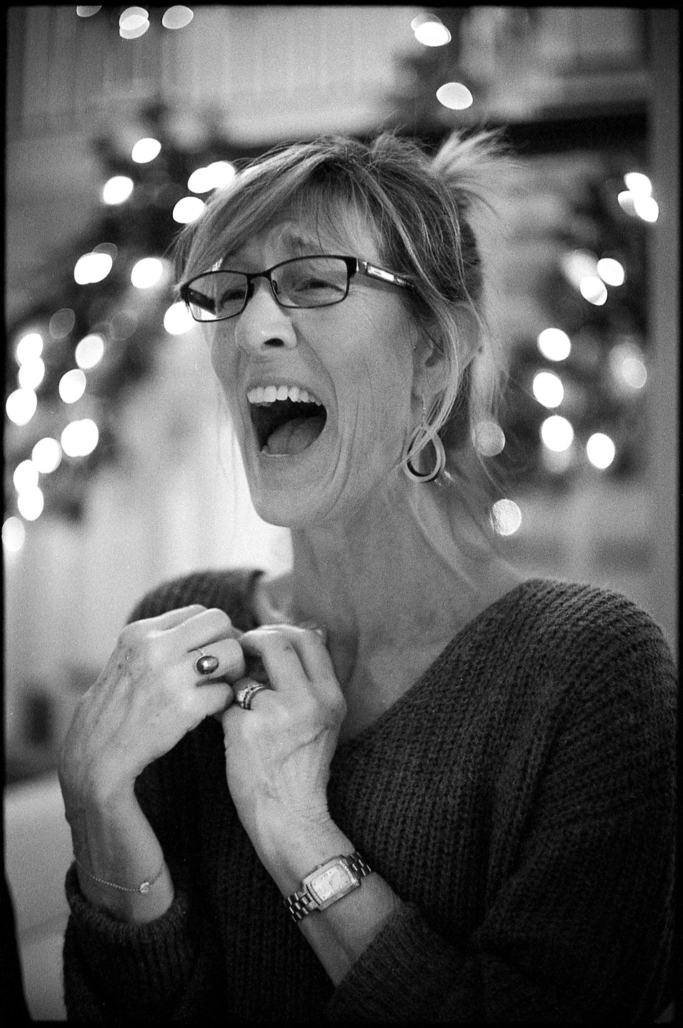 A woman with glasses laughs with her mouth wide open, holding her hands to her chest, with bokeh lights in the background.