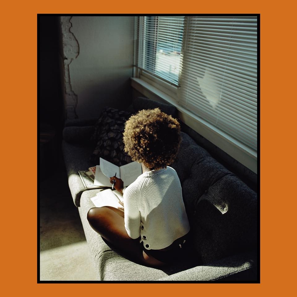 A woman with an afro sits on a sofa, writing in a notebook by a window with blinds.