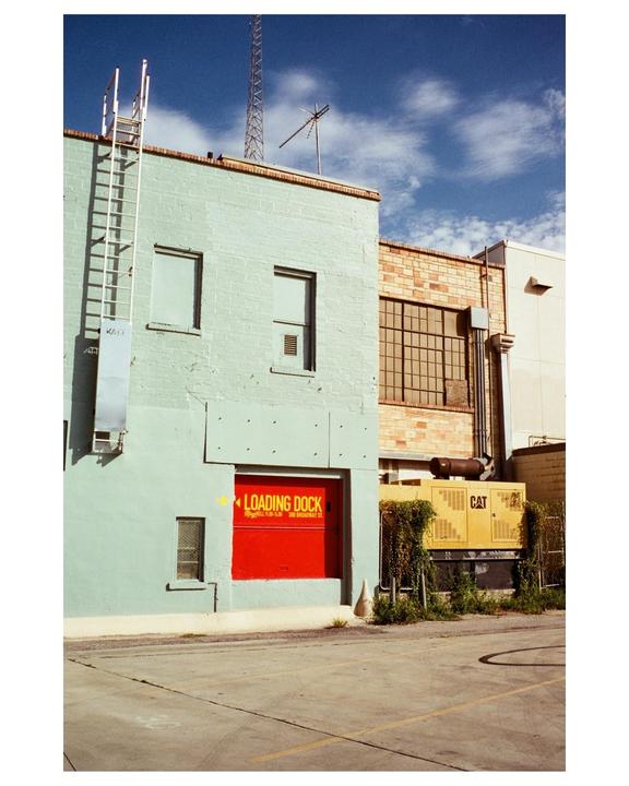 A light blue building with a red loading dock door stands next to a brick building under a blue sky with clouds.
