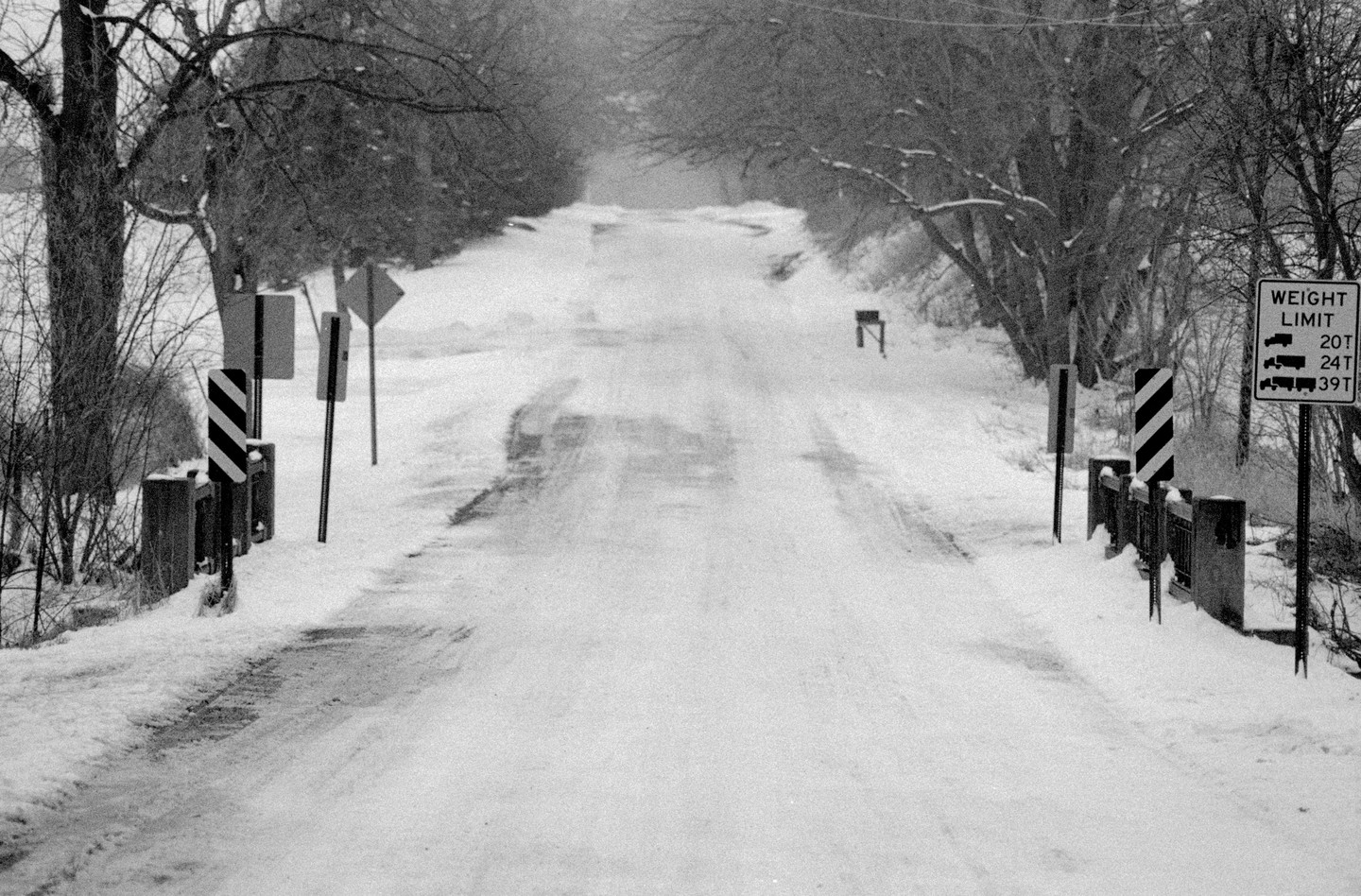A snow-covered road with a weight limit sign on the right leads into the distance.