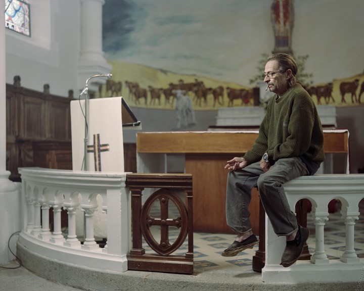 A man sits on a white railing in a church, with a mural of cattle in the background.
