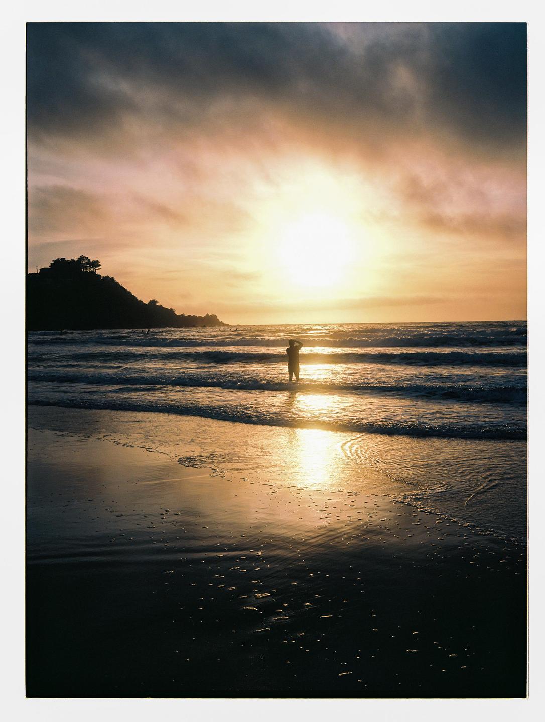 A person stands in the ocean at sunset with a silhouetted hill on the left.