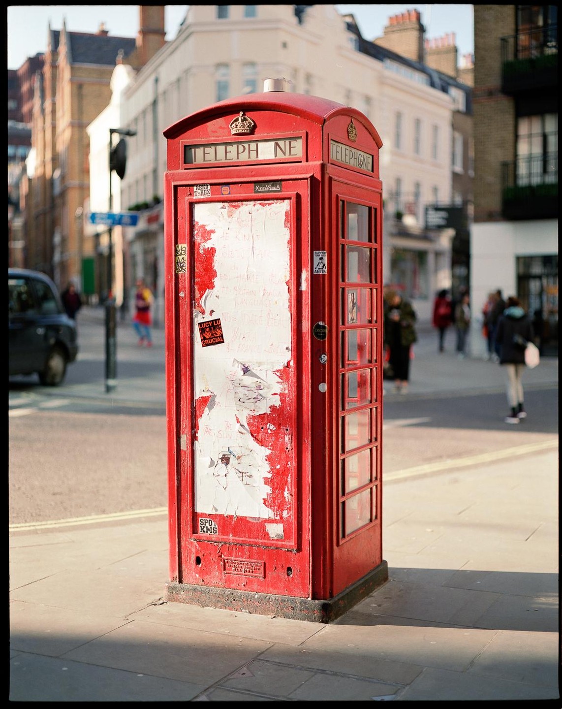 A red telephone booth stands on a city street with buildings and people in the background.