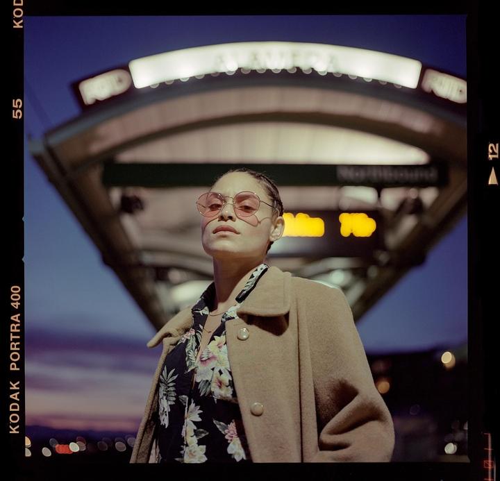 A woman wearing a floral shirt, a coat, and pink sunglasses stands under a sign at dusk.