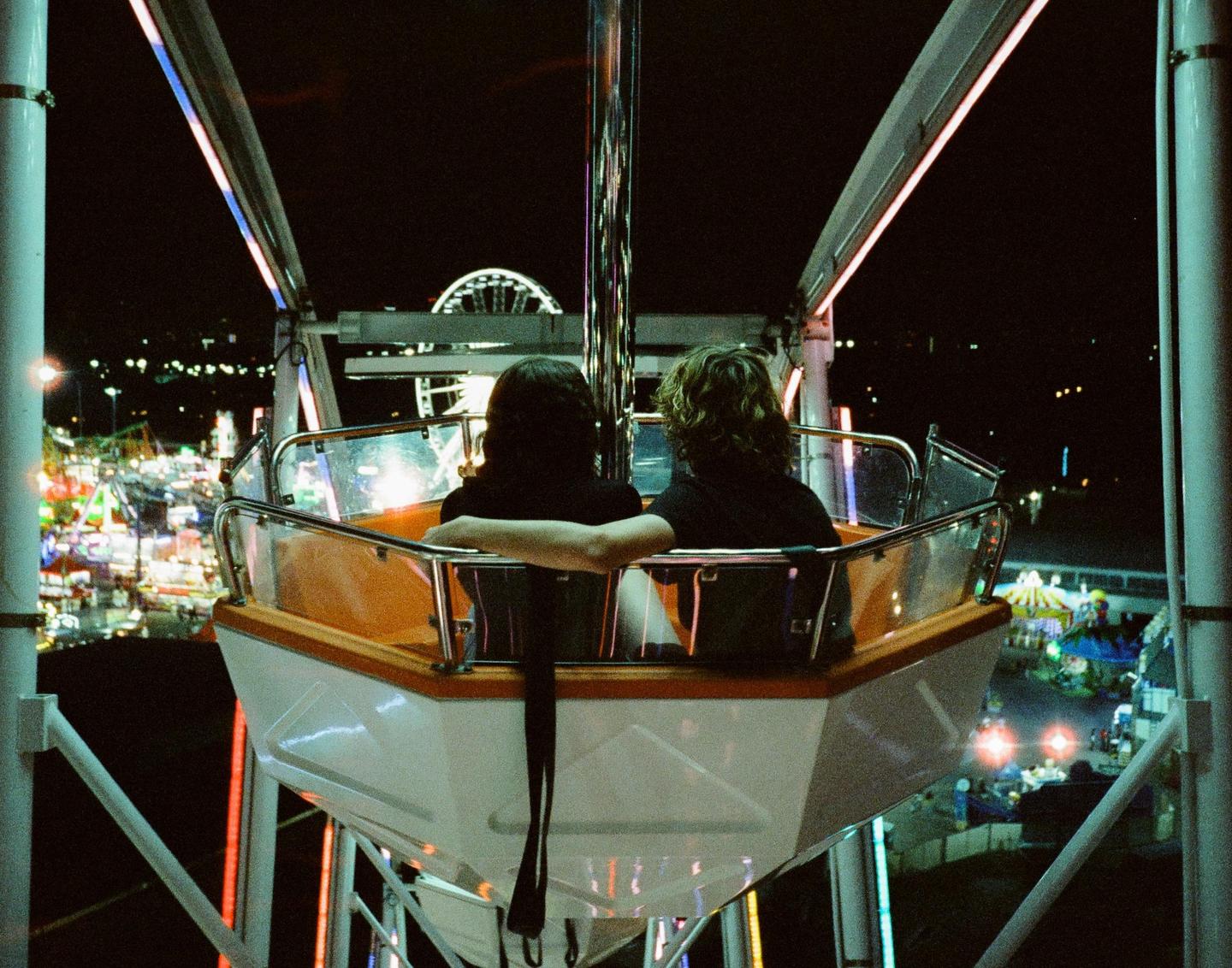 Two people sit in a ferris wheel gondola overlooking a fairground at night.