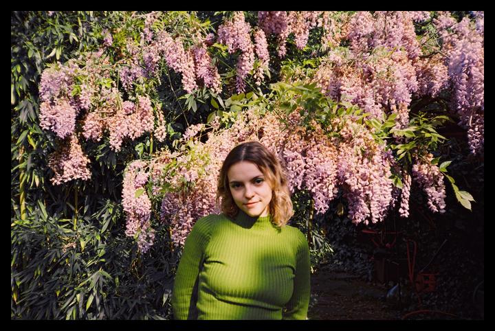 A woman in a green sweater stands in front of a wall of wisteria blossoms.