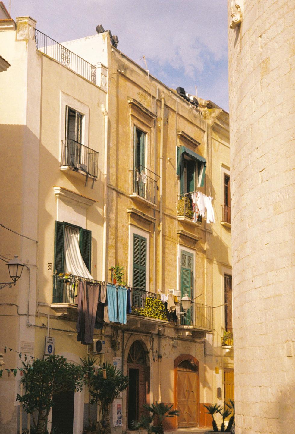Laundry hangs from balconies on a street with tall buildings and a stone column.