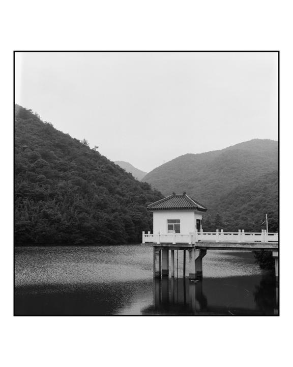 A small building with a tiled roof stands on stilts over a lake, connected to the shore by a bridge, with forested mountains in the background.
