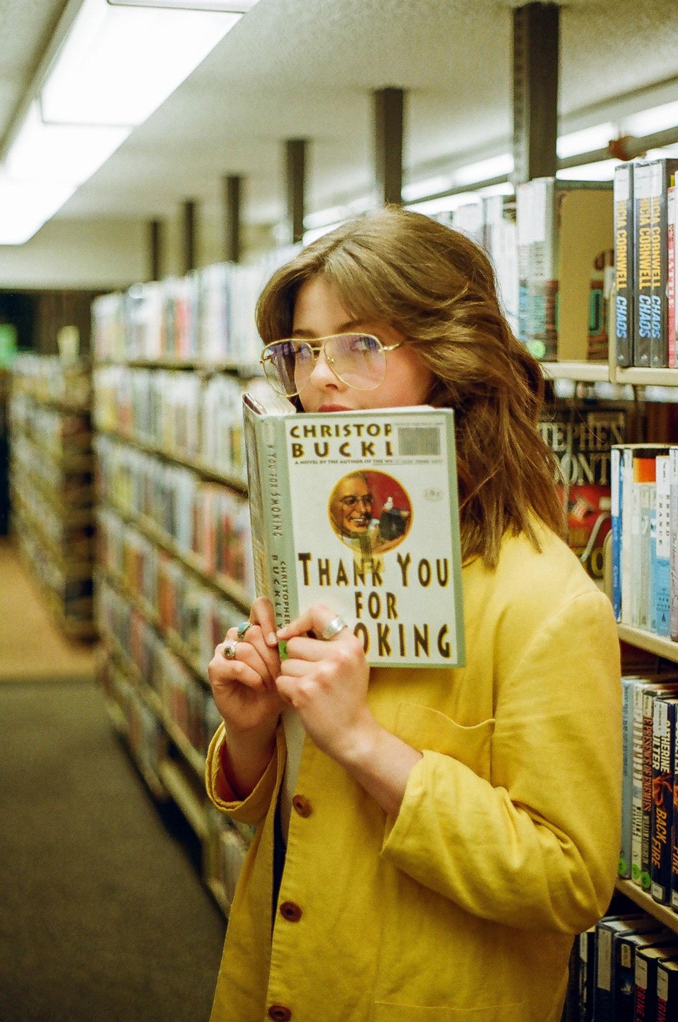 A woman in a yellow jacket holds a book in front of her face in a library.