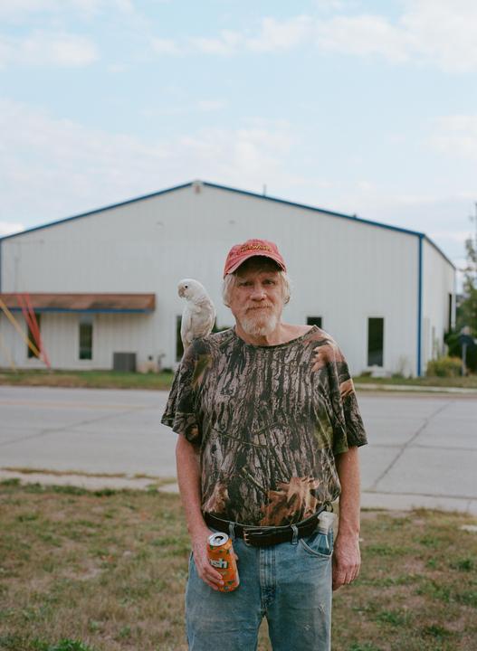 A man with a beard and a parrot on his shoulder stands outdoors holding a soda can.