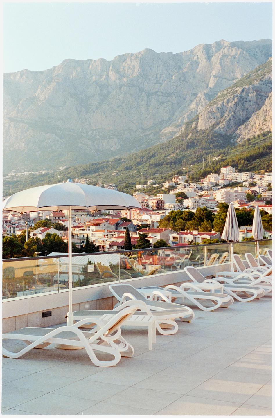 A row of lounge chairs and umbrellas on a patio overlooks a town nestled at the base of a large mountain.