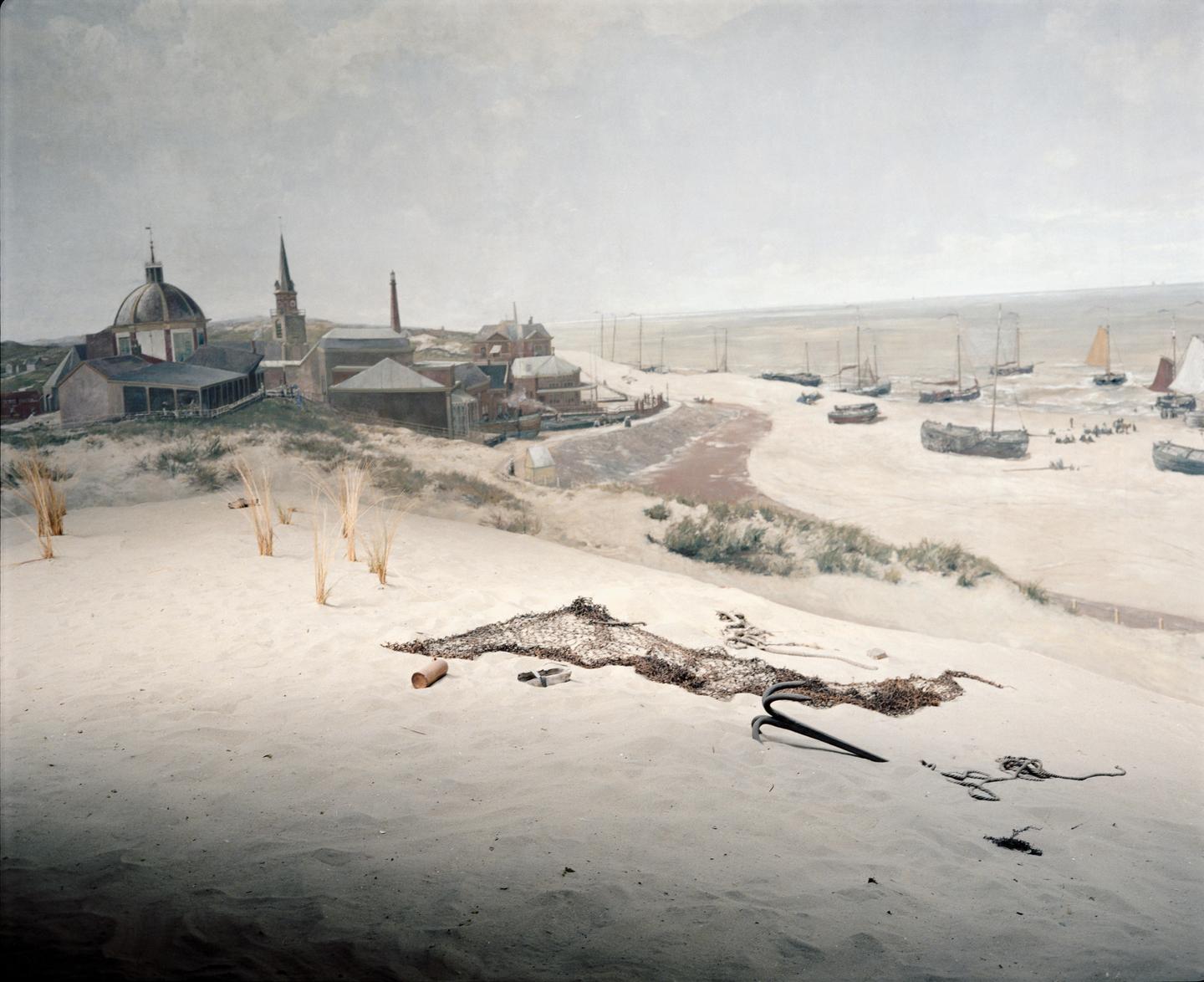 A fishing village with boats on the sand and in the water is seen from a sandy dune with a fishing net and anchor in the foreground.