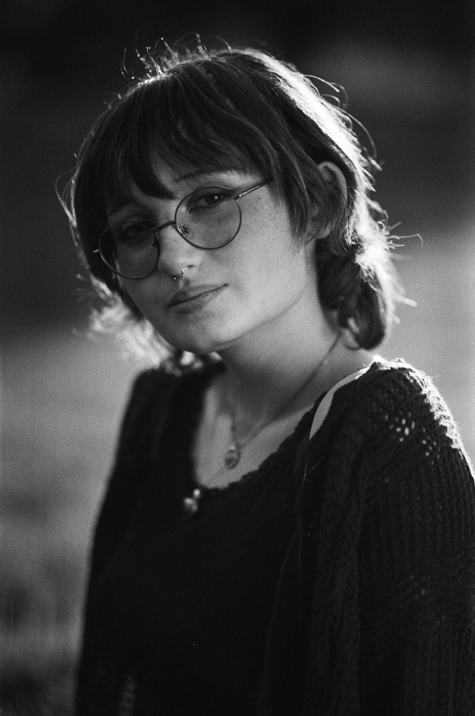A young woman with glasses and a nose ring looks towards the viewer.