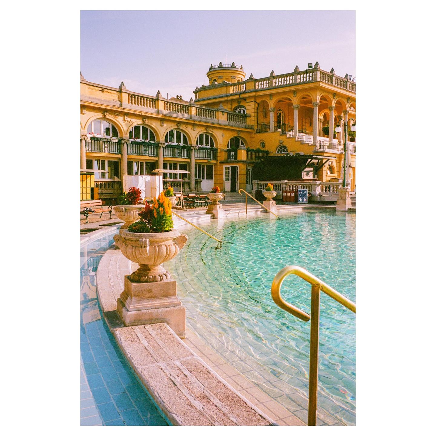 A large yellow building with arched windows overlooks a swimming pool with clear blue water and a golden handrail.