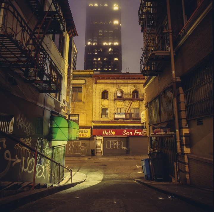 A narrow street is illuminated by a street light and the glowing windows of a tall building in the background.