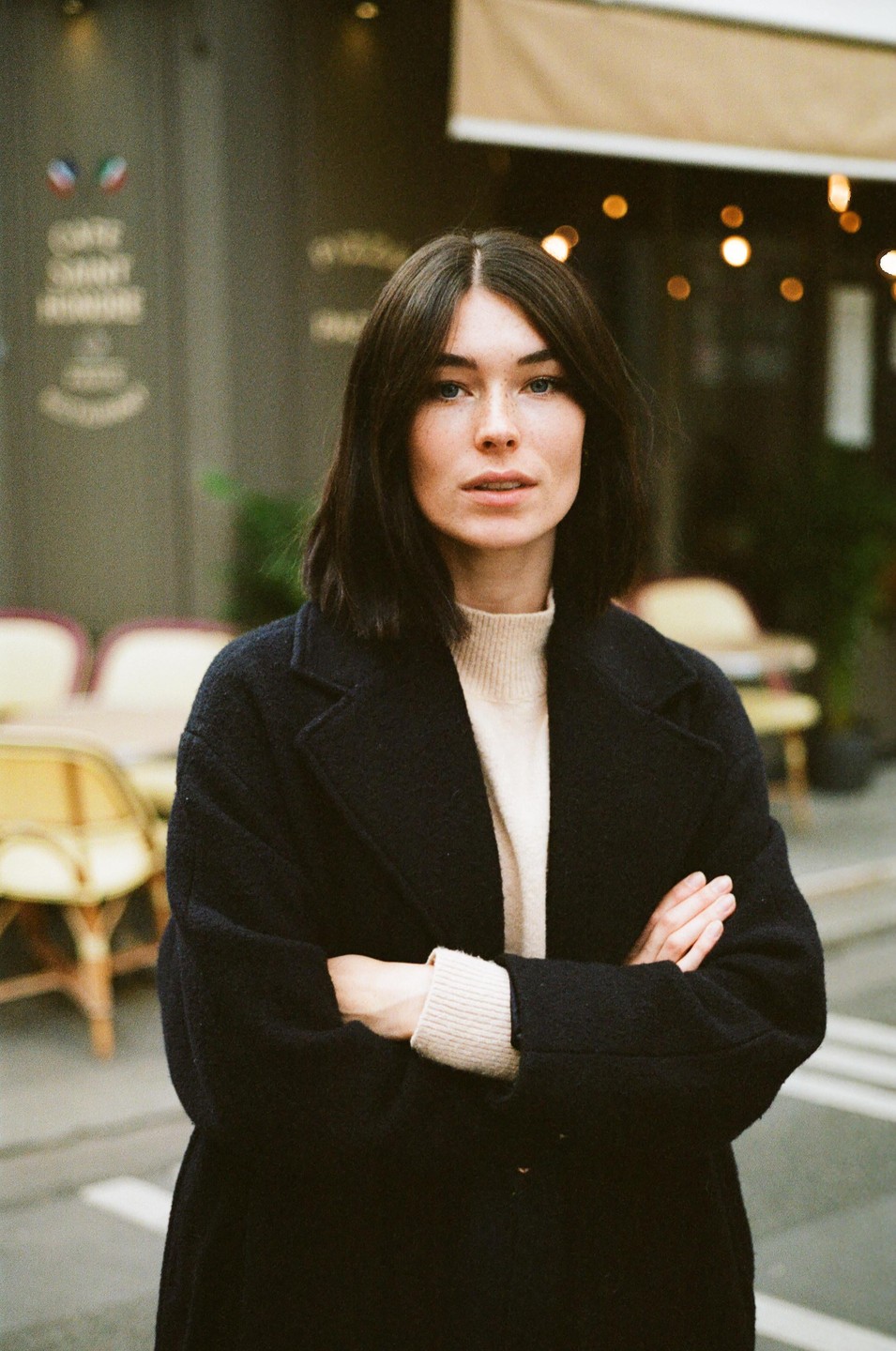 A woman with dark hair and a black coat stands with her arms crossed in front of a cafe.