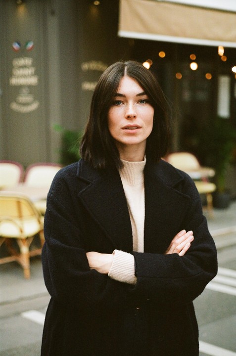 A woman with dark hair and a black coat stands with her arms crossed in front of a cafe.