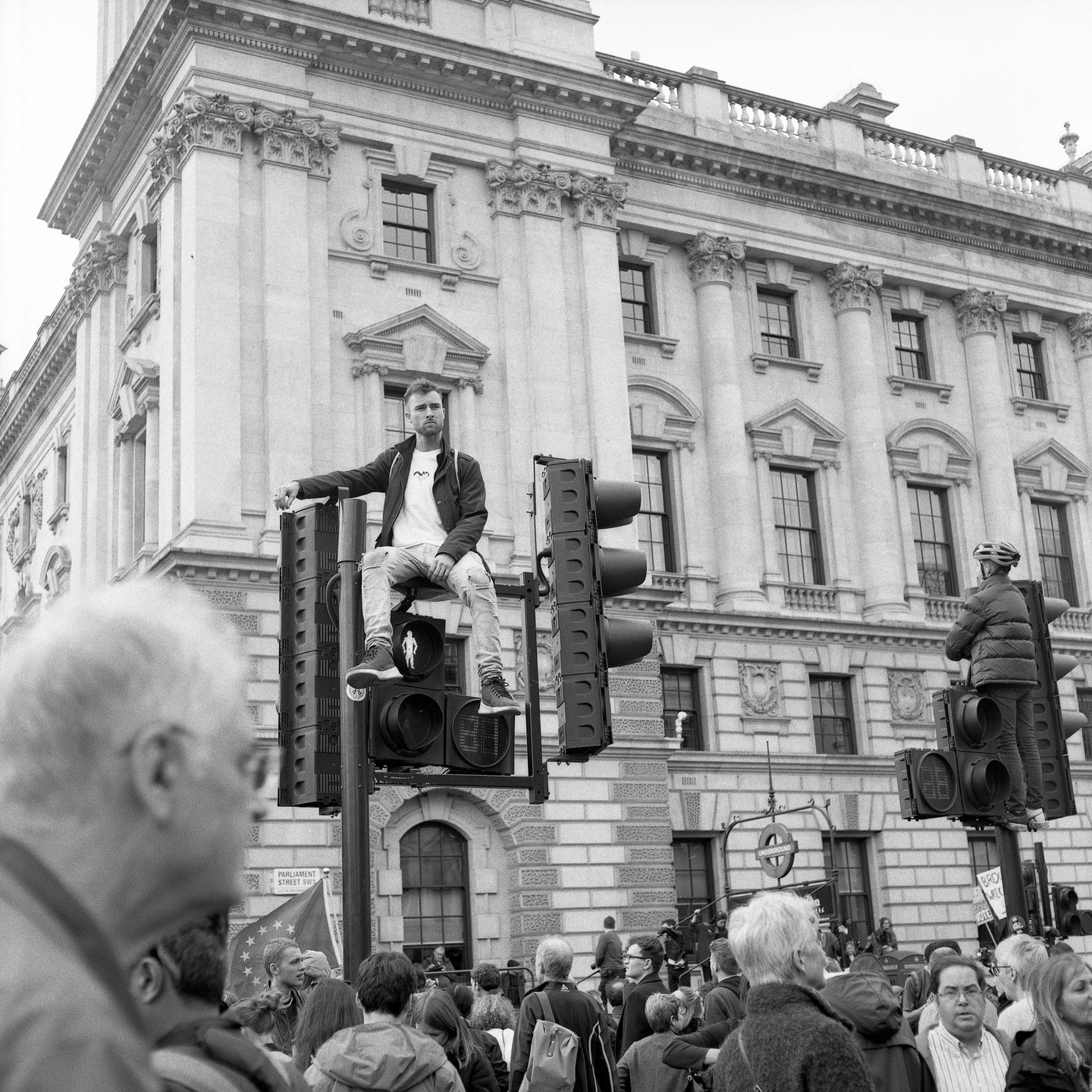 A man sits atop a traffic light pole while another man stands on a different traffic light pole, both overlooking a crowd of people on a street in front of a large building.