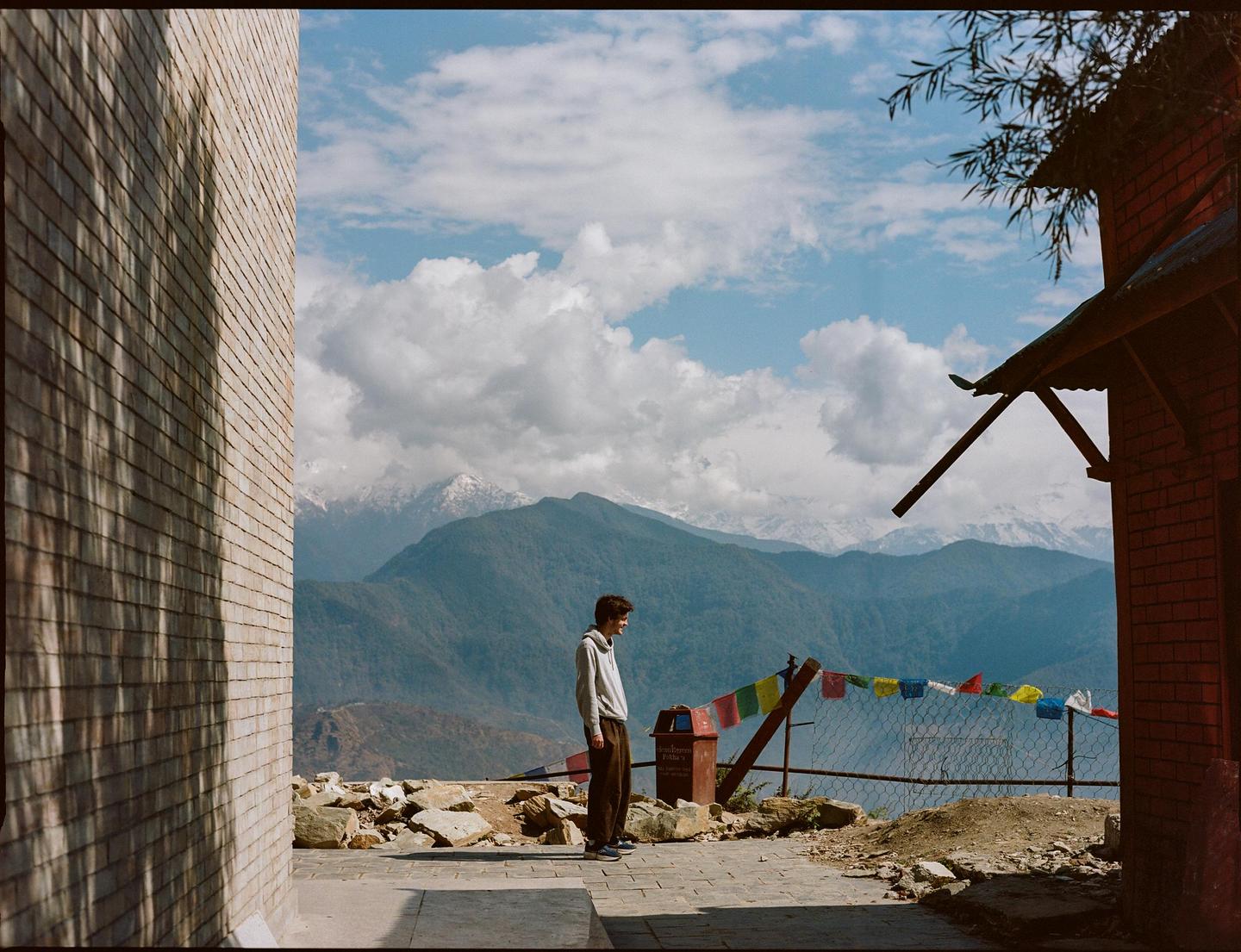 A man stands on a paved area overlooking a mountain range with prayer flags strung along a fence.