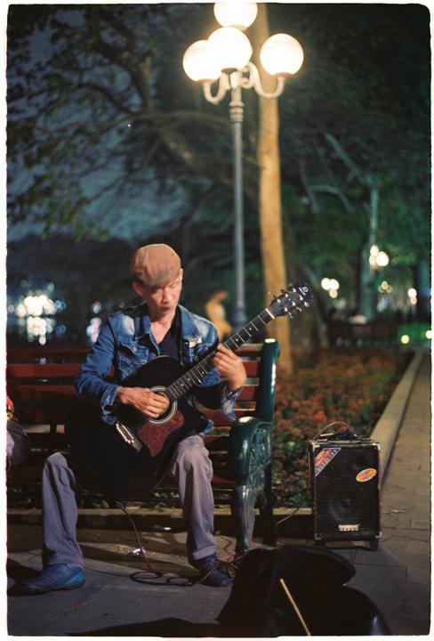 A man wearing a denim jacket and a cap sits on a park bench playing an acoustic guitar at night.