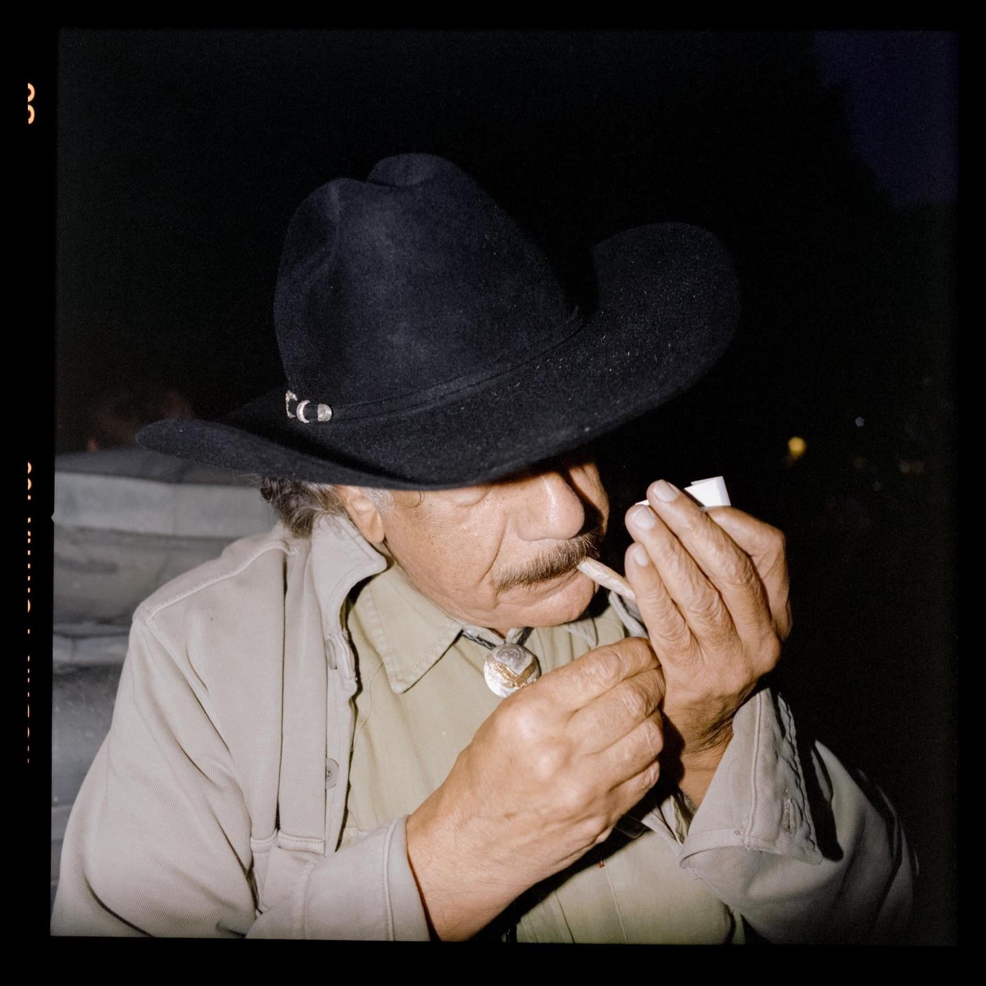 A man wearing a cowboy hat lights a cigarette at night.