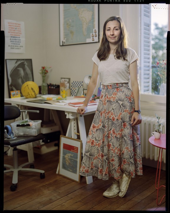 A woman stands in a room with a desk, a map on the wall, and a window with shutters.