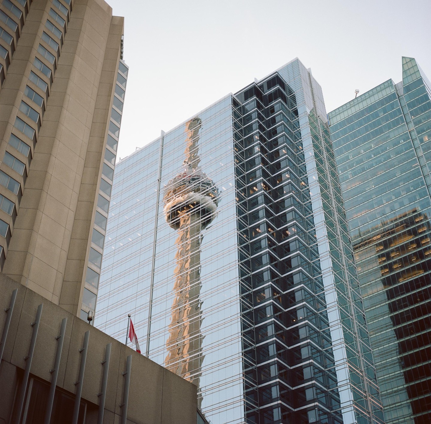 The CN Tower is reflected in the glass facade of a modern skyscraper.