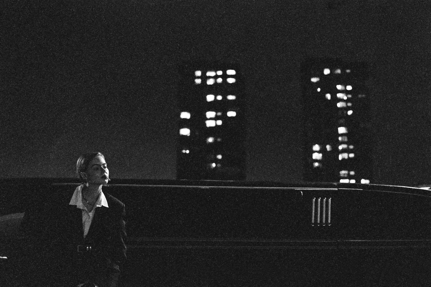 A woman with a cigarette in her mouth stands next to a limousine at night with city lights in the background.