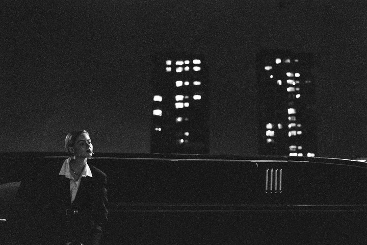 A woman with a cigarette in her mouth stands next to a limousine at night with city lights in the background.