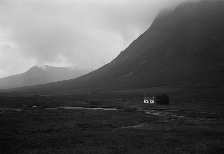 A small white house sits in a vast valley with mountains shrouded in fog.
