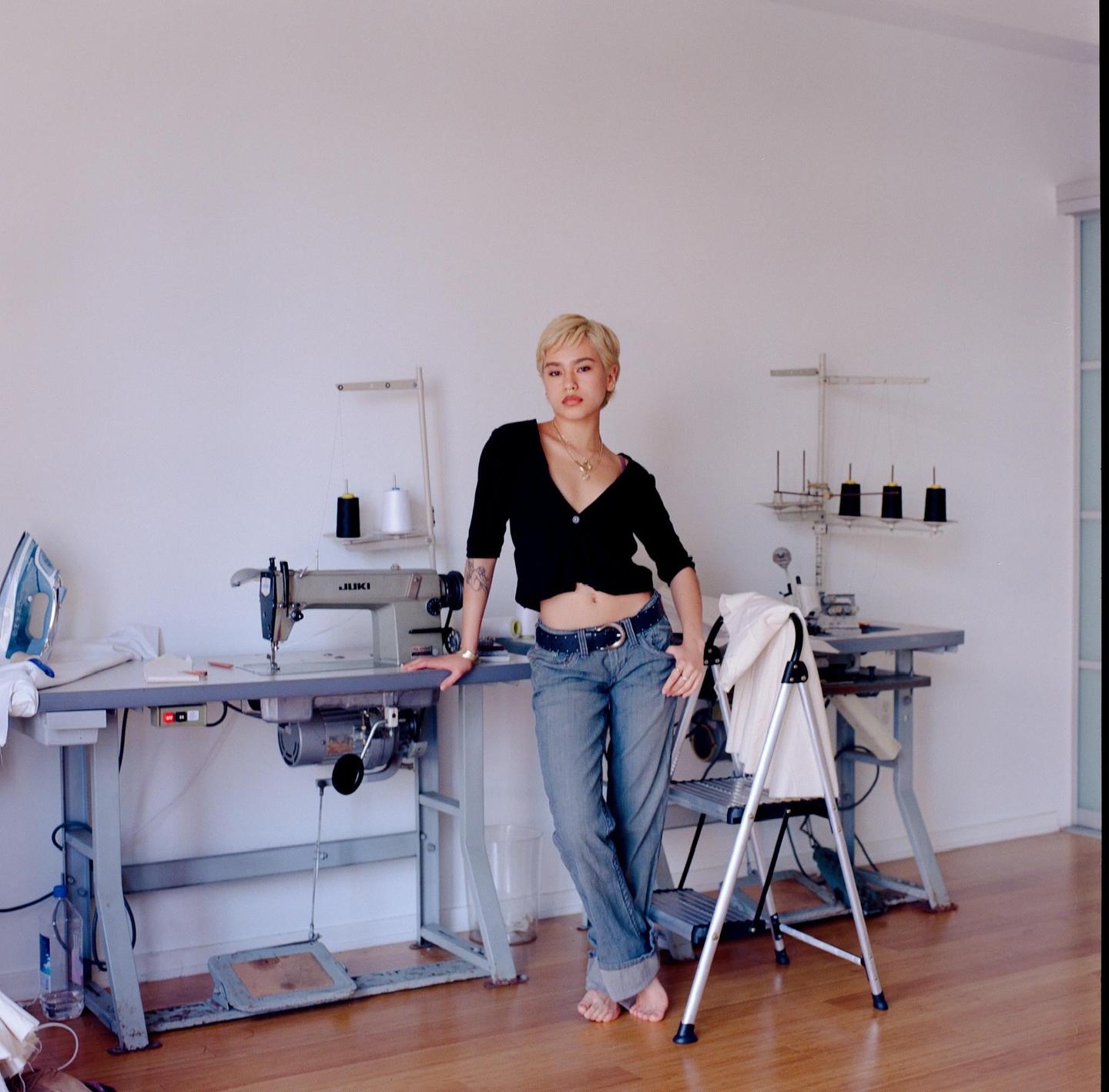 A woman with short blonde hair stands barefoot in a studio, leaning against a sewing machine.