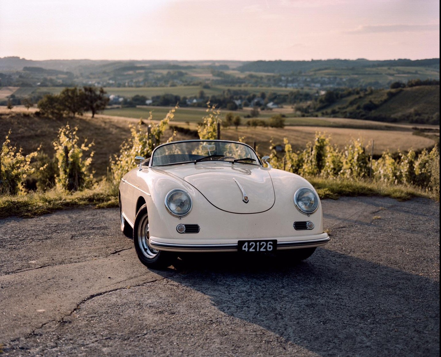 A cream-colored vintage convertible car is parked on a paved road overlooking a valley with vineyards and a village in the distance.