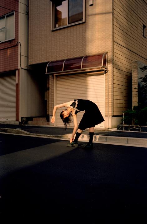A woman in a black dress bends over to tie her shoe on a street with buildings in the background.