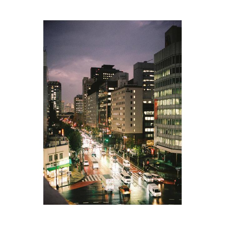 A city street is filled with cars and buildings under a cloudy sky at dusk.