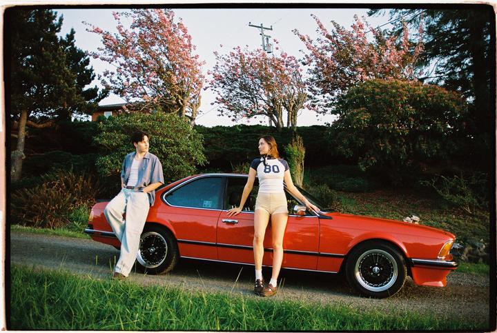 A man and a woman pose with a red car on a grassy roadside next to flowering trees.