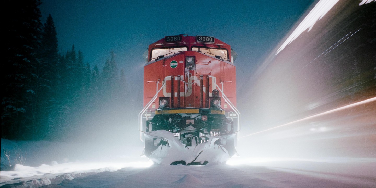 A red train with bright headlights moves through a snowy landscape with a forest in the background.