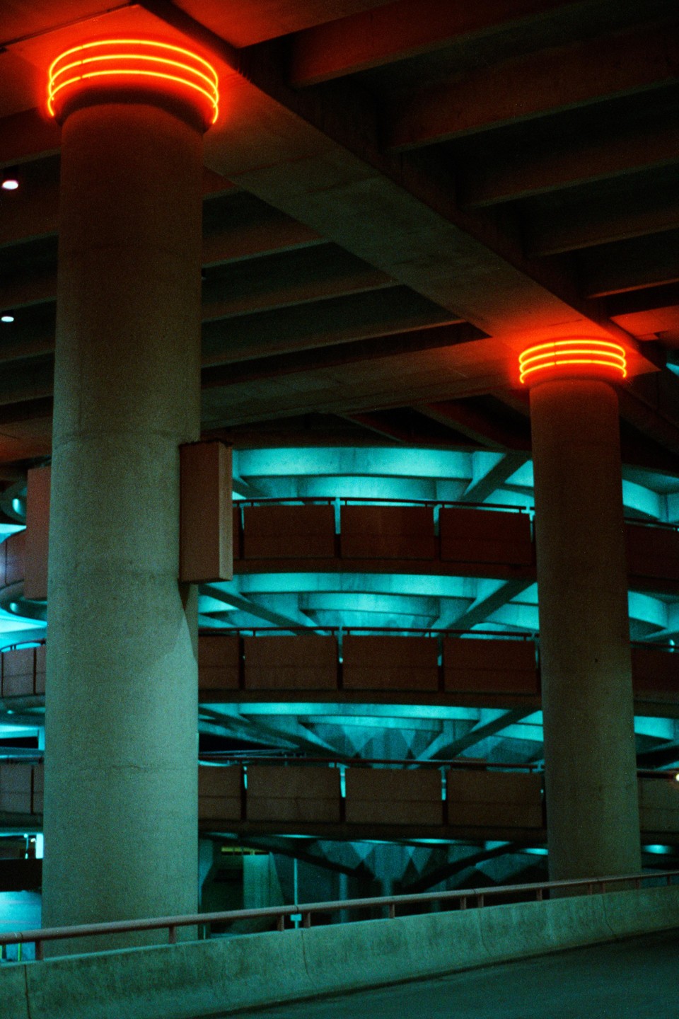 Two concrete pillars with orange neon lights on top stand in a parking garage with teal lighting.