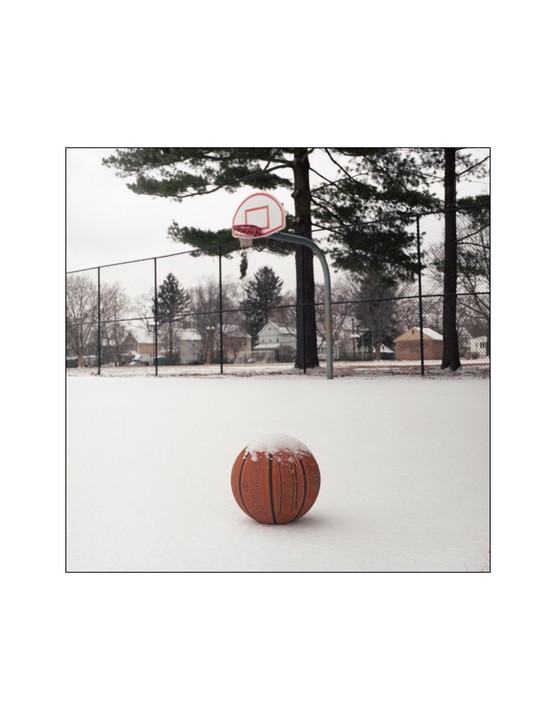 A basketball covered in snow sits on a snow-covered court with a basketball hoop and trees in the background.