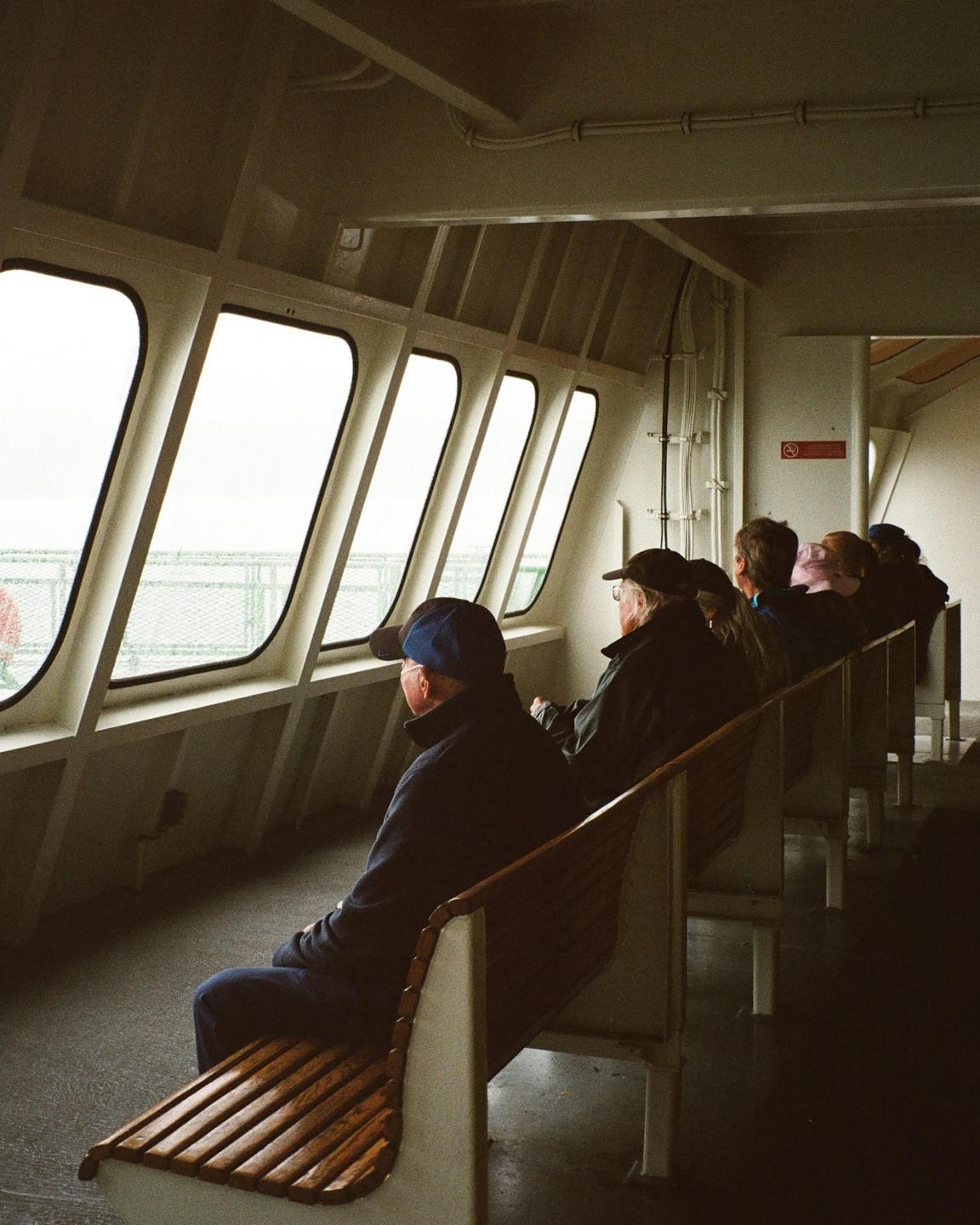 People sit on wooden benches inside a ferry, looking out of large windows.