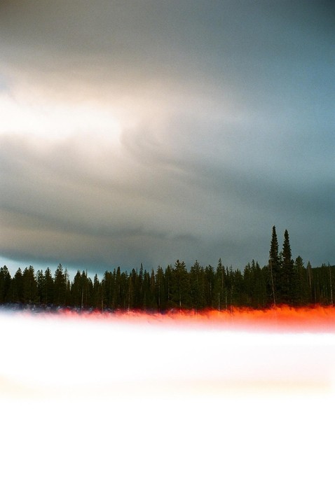 A dark forest stands under a cloudy sky with a bright orange light leak across the bottom.