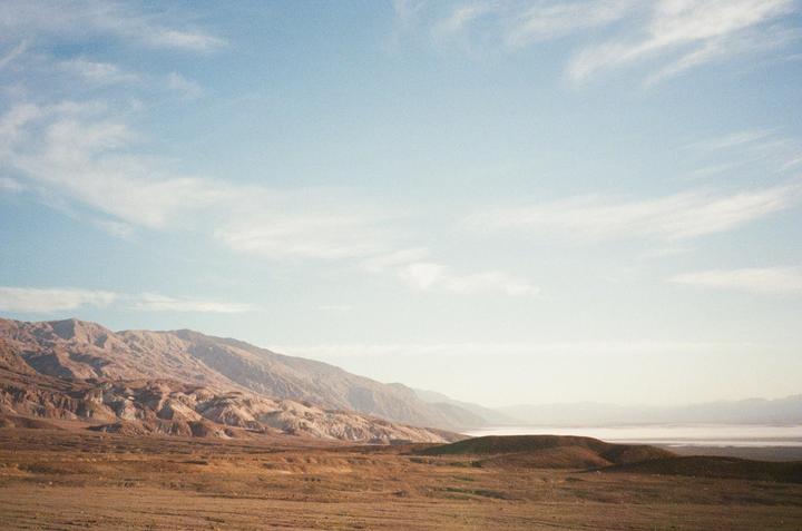A vast desert landscape features mountains, hills, and a distant body of water under a light blue sky with wispy clouds.