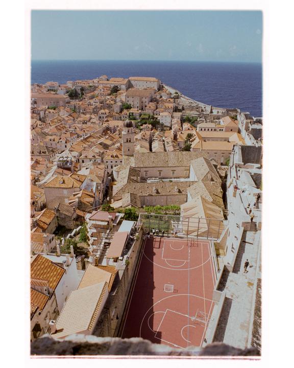An aerial view shows a red basketball court nestled among buildings with terracotta roofs, with the sea in the background.
