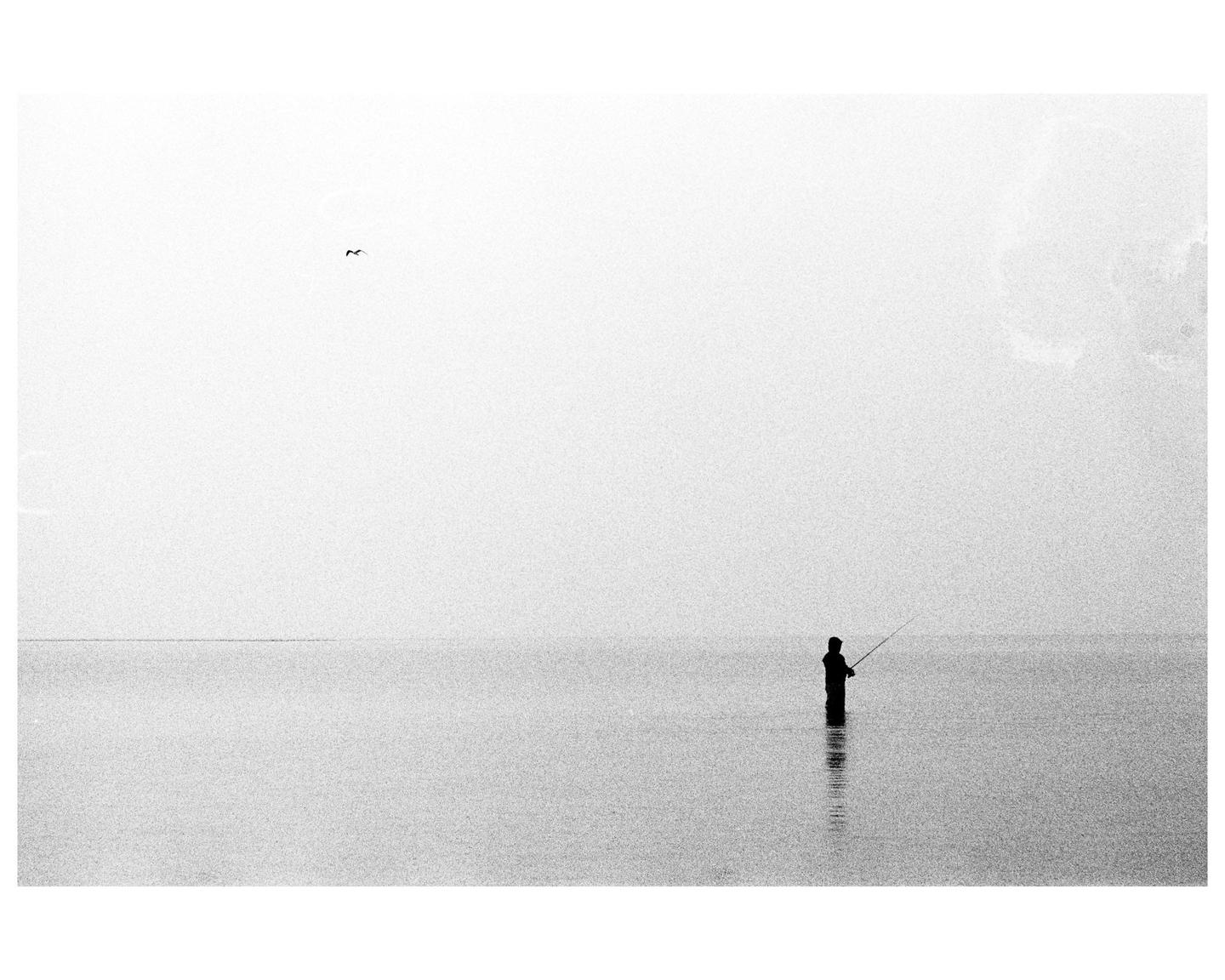 A lone fisherman stands in the water with a fishing rod under a hazy sky.