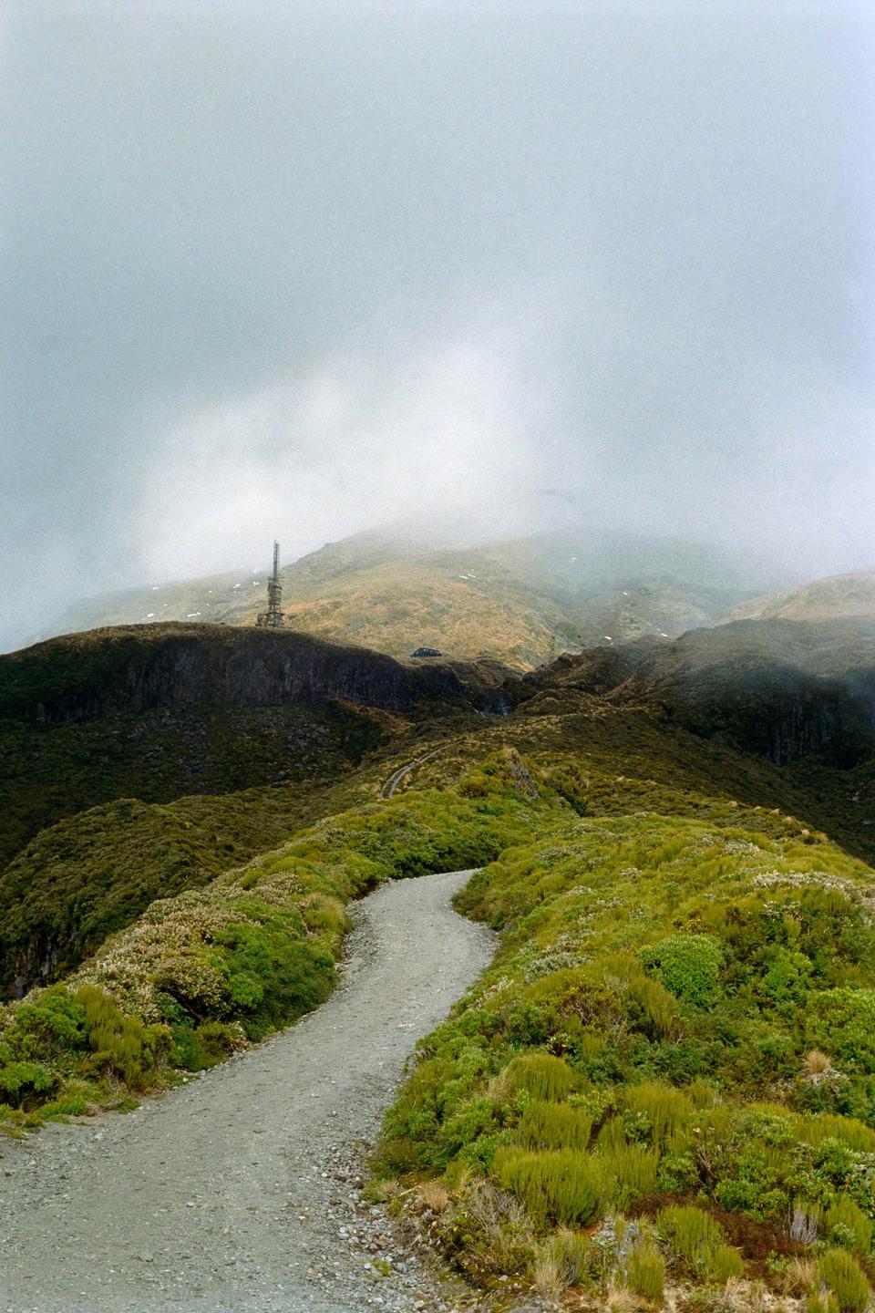 A winding path leads through a green, mountainous landscape with a communication tower visible in the distance under a cloudy sky.